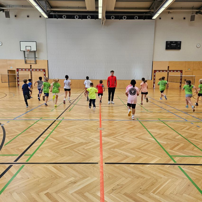 Eine Gruppe von Kindern spielt ein Spiel in einer Turnhalle. Ein Trainer beobachtet sie. Die Turnhalle hat Holzböden und Wände.