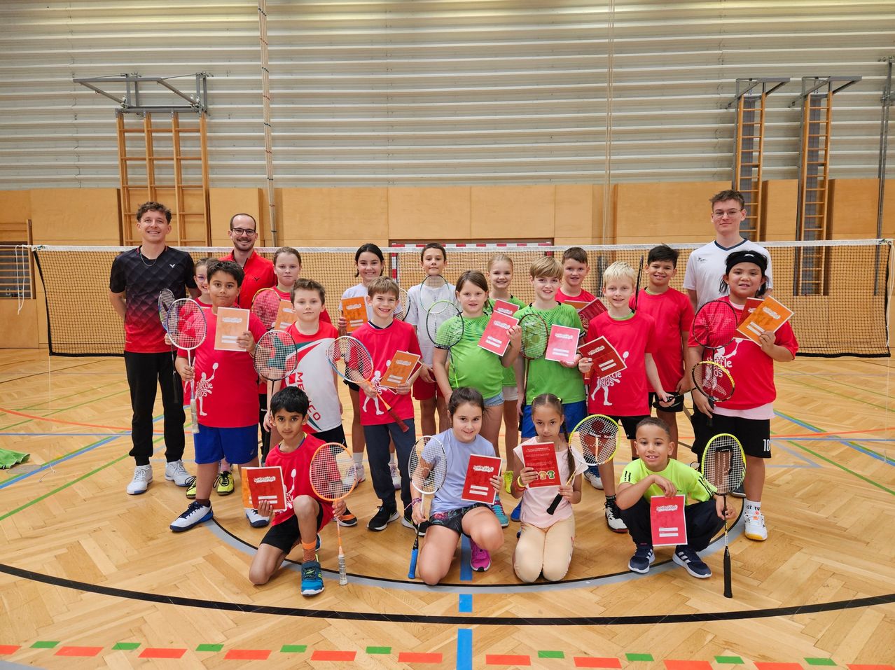 Eine Gruppe von Kindern posiert mit Badmintonschlägern und hält Urkunden in der Hand, in einer Turnhalle mit Holzboden und einem Netz im Hintergrund.