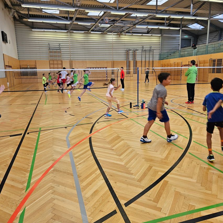 Eine Gruppe von Kindern spielt Badminton drinnen. Sie sind in Teams aufgeteilt und rennen über das Spielfeld.