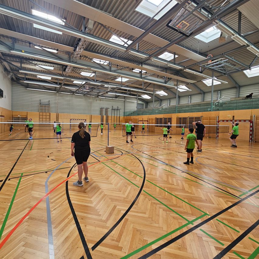 Eine Sporthalle mit Holzboden hat mehrere Volleyballnetze aufgestellt. Eine Gruppe von Menschen in grünen Trikots spielt. Eine Frau in Schwarz steht in der Nähe des mittleren Netzes.
