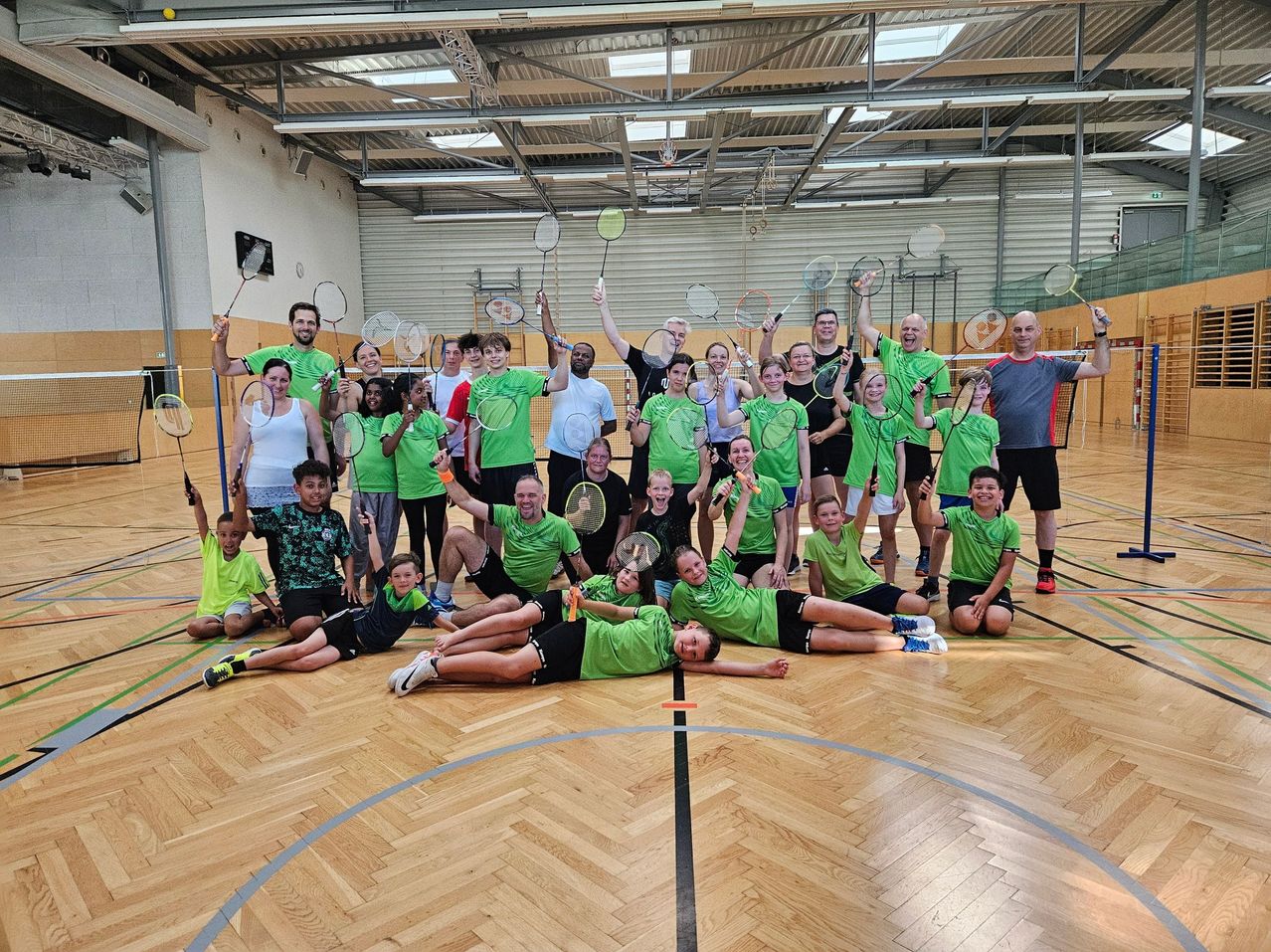 Eine Gruppe von Menschen in grünen Uniformen posiert für ein Foto auf einem Basketballplatz, wobei sie Badmintonschläger halten. Der Platz hat Holzboden und blaue Linien.