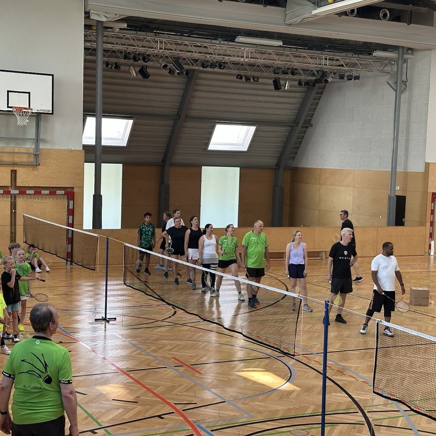 In einer Indoor-Sporthalle stehen sich mehrere Personen für ein Badmintonspiel gegenüber. Sie halten Schläger in der Hand und warten auf den Beginn des Spiels.