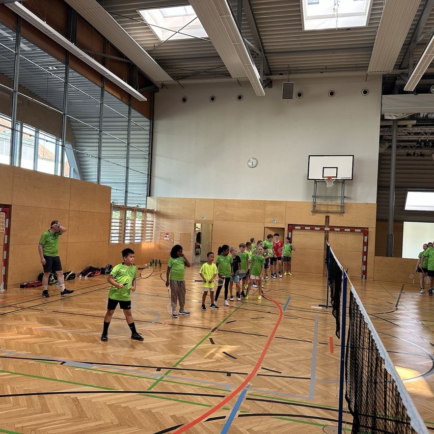 Eine Gruppe von Kindern in grünen Shirts und Shorts spielt Badminton in einer Halle mit Holzböden. Eine Person steht an der Seite.
