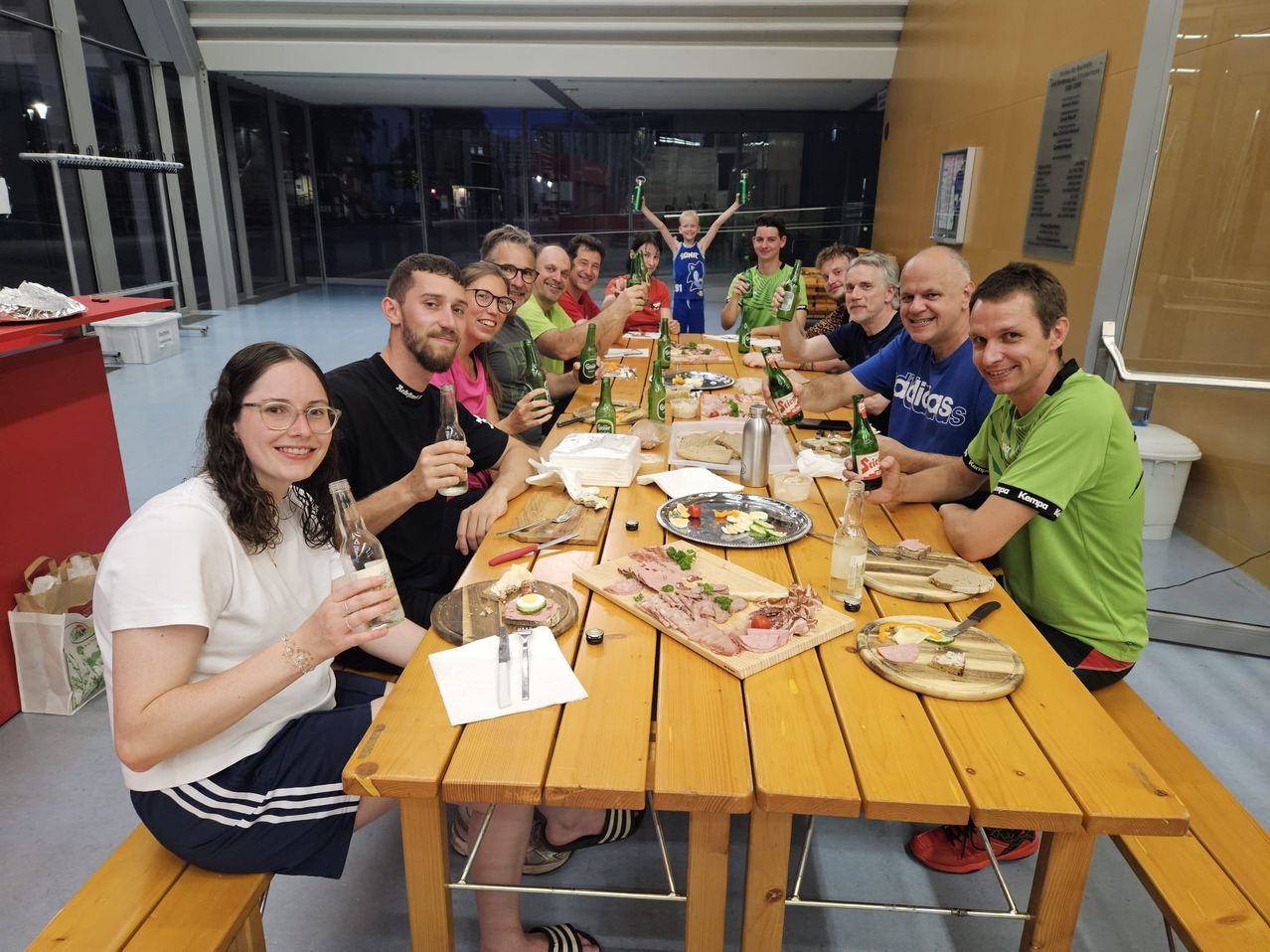 Eine Gruppe von Menschen sitzt um einen Tisch im Innenbereich, lächelt und hält Flaschen. Auf dem Tisch stehen Teller mit Essen, einschließlich Fleisch und Brot. Ein Kind steht hinter der Gruppe und hält eine Flasche in die Luft.
