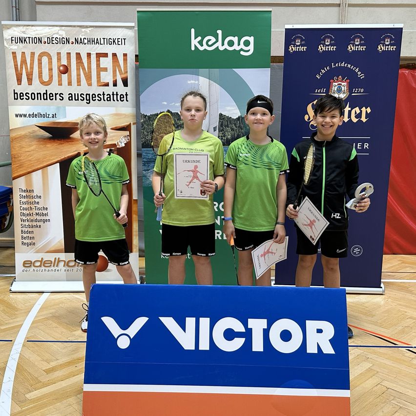 Vier junge Jungen stehen in einer Turnhalle, halten Badmintonschläger und Urkunden. Sie tragen grüne und schwarze Trikots. Hinter ihnen stehen drei Banner, von denen eines Kelag sagt.