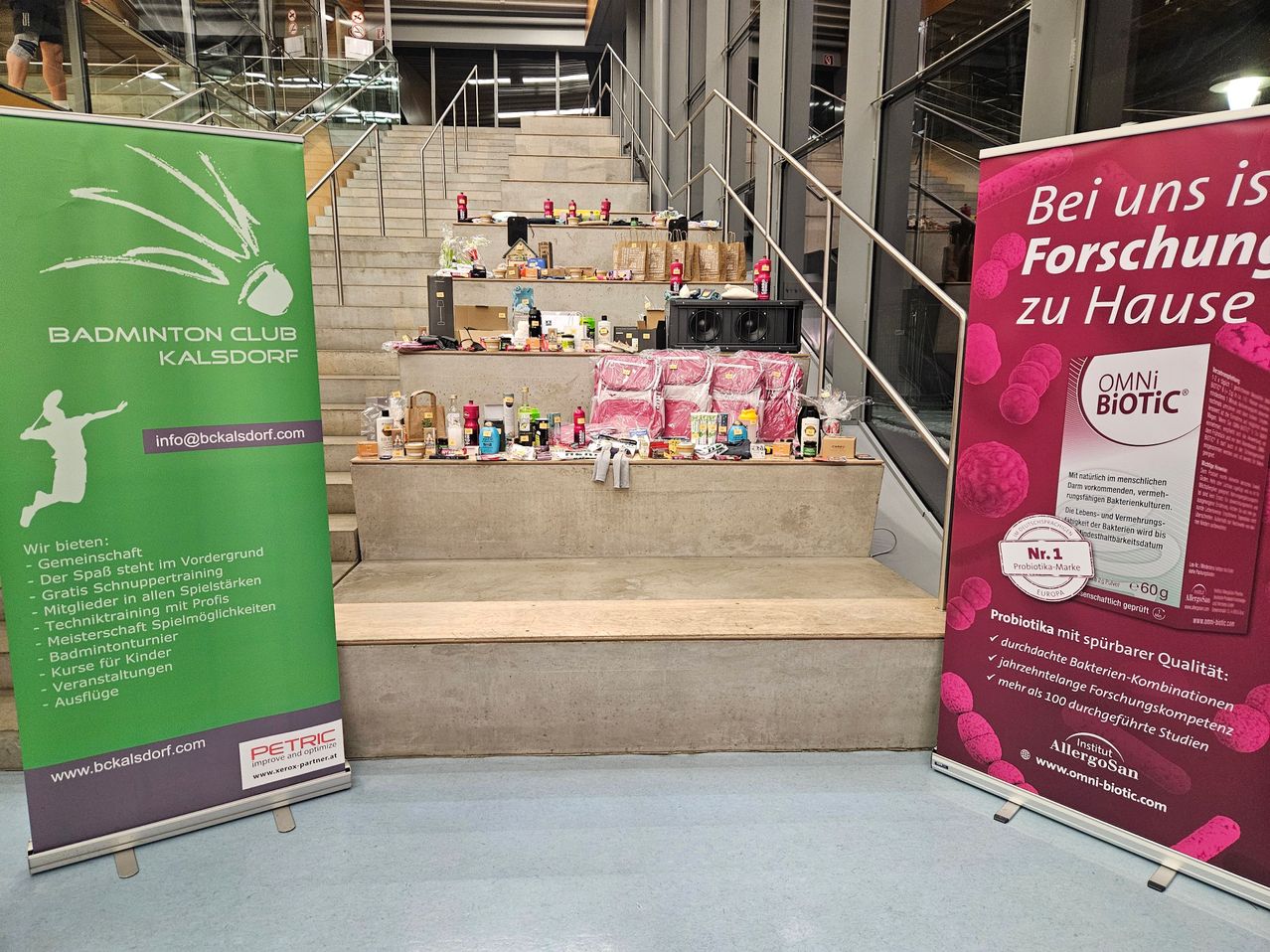 Eine Innentreppe mit zahlreichen Geschenktüten und Produkten, die auf ihren Stufen ausgestellt sind. Zwei Banner, eines grün und eines rosa, stehen neben der Treppe. Das grüne Banner hat weißen Text und ein Logo, während das rosa Banner roten und weißen Text mit einem Logo hat.