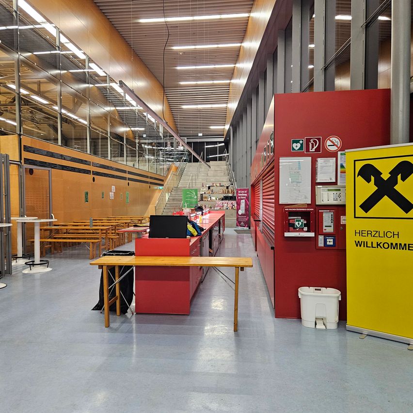 Ein leerer Raum mit Tischen, Bänken und einer roten Theke. Der Raum hat eine hohe Decke mit Lichtern und Glaswänden. Es gibt eine Treppe auf der linken Seite. Rechts befindet sich ein gelbes Schild mit dem Wort 'Herzlich willkommen'.