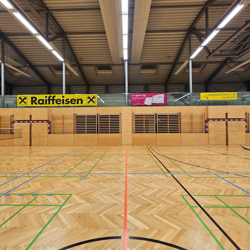 Eine leere Indoor-Sporthalle mit Holzboden, mehreren Türen und Bannern. Ein Banner sagt 'Raiffeisen'. Ein weiteres Banner sagt 'Ein guter Tag beginnt mit'. Die Wände sind aus Holz mit einigen Geräten.