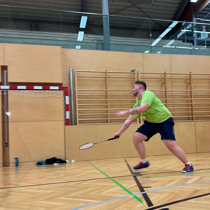 Ein Mann spielt Badminton in einer Halle. Er ist in Bewegung und schlägt einen Shuttlecock. Er trägt ein grünes Shirt und dunkle Shorts. Der Platz hat Holzböden und blaue Markierungen.