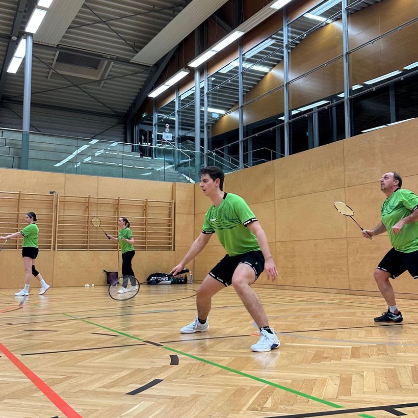 Fünf Personen spielen Badminton in einer Turnhalle. Einer ist bereit, den Ball zu schlagen. Ein anderer konzentriert sich auf den Ball. Zwei weitere sind an den Seitenlinien.