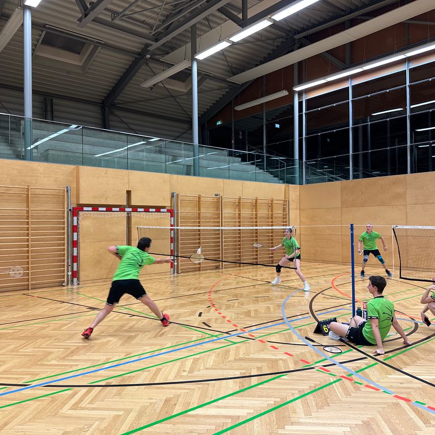 In einer Halle spielen eine Gruppe von Spielern Badminton. Sie tragen grüne Shirts und schwarze Shorts. Alle halten Badmintonschläger in den Händen.