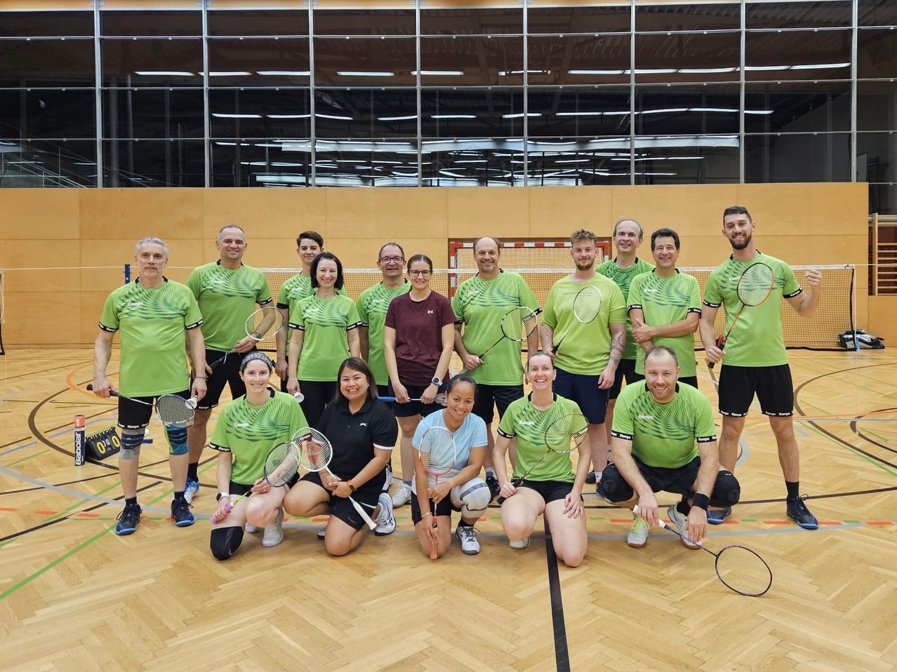 Eine Gruppe von Menschen in grünen Shirts und mit Badmintonschlägern posiert für ein Foto in einer Sporthalle.