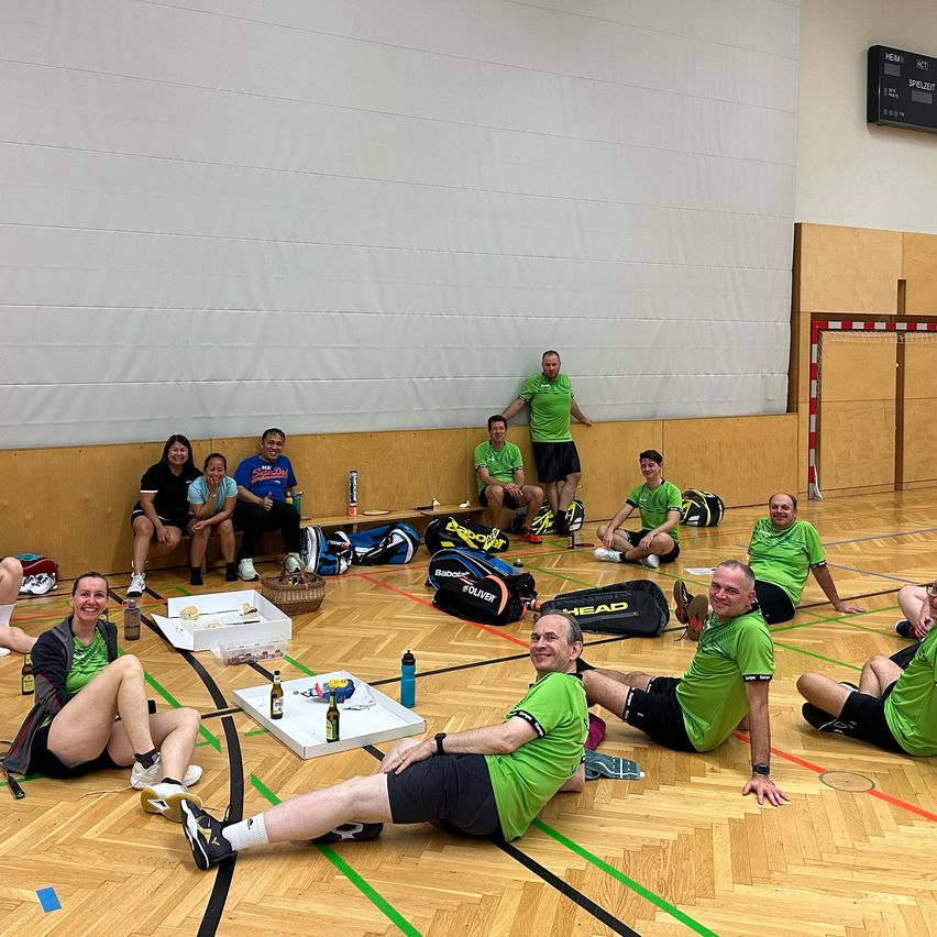 Eine Gruppe von Menschen in grünen Shirts sitzt auf dem Boden einer Turnhalle, einige entspannen sich, während andere einen Tisch mit Essen und Getränken betrachten.