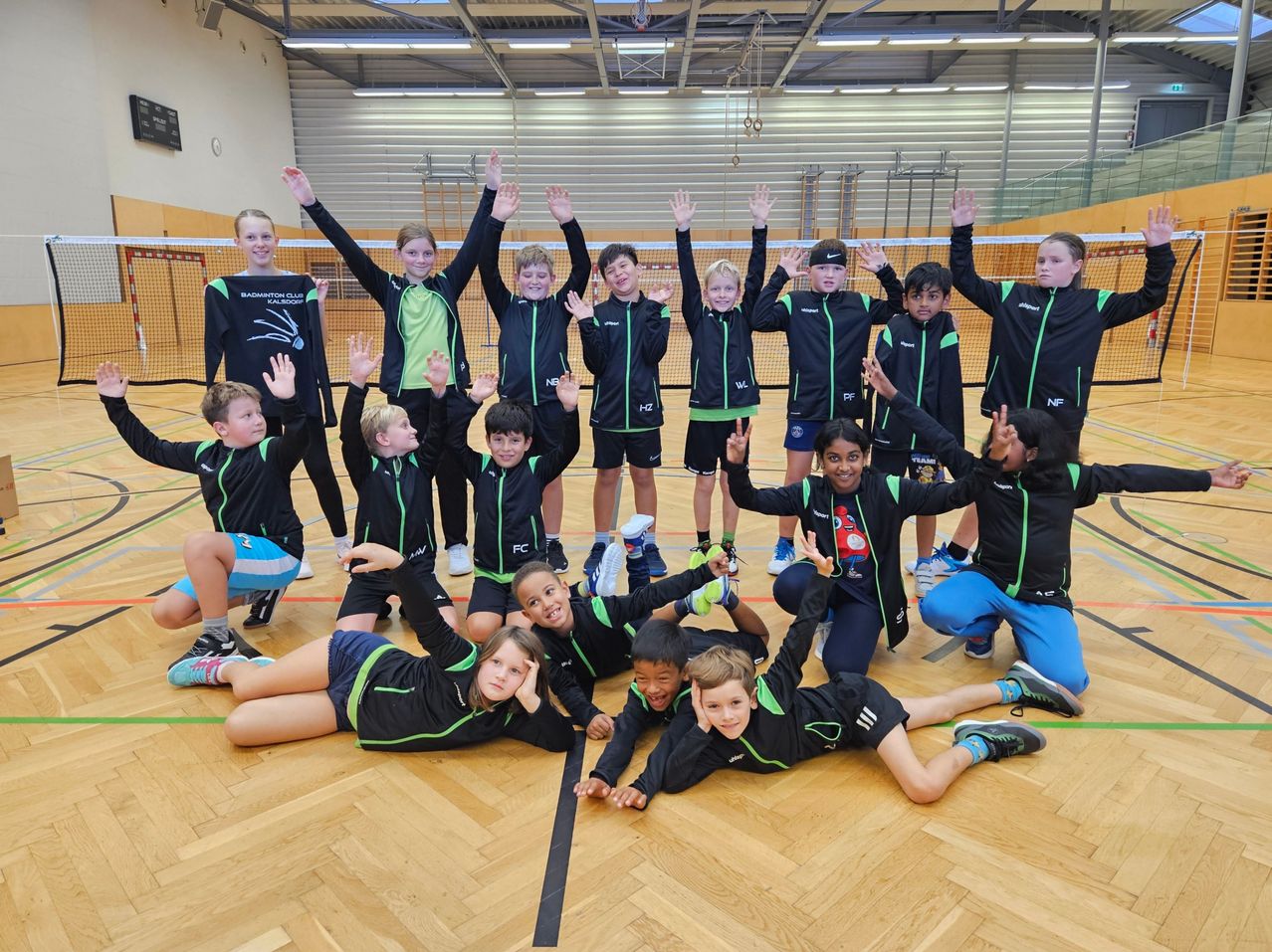 Eine Gruppe junger Spieler in schwarzen und grünen Sportuniformen posiert für ein Foto in einer Turnhalle, einige liegen auf dem Boden und andere stehen hinter einem Netz.