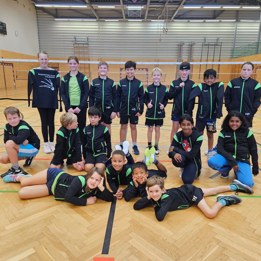 Eine Gruppe junger Athleten in Sportbekleidung posiert für ein Foto in einer Turnhalle. Sie liegen auf dem Boden, einige stehen dahinter, und alle lächeln.