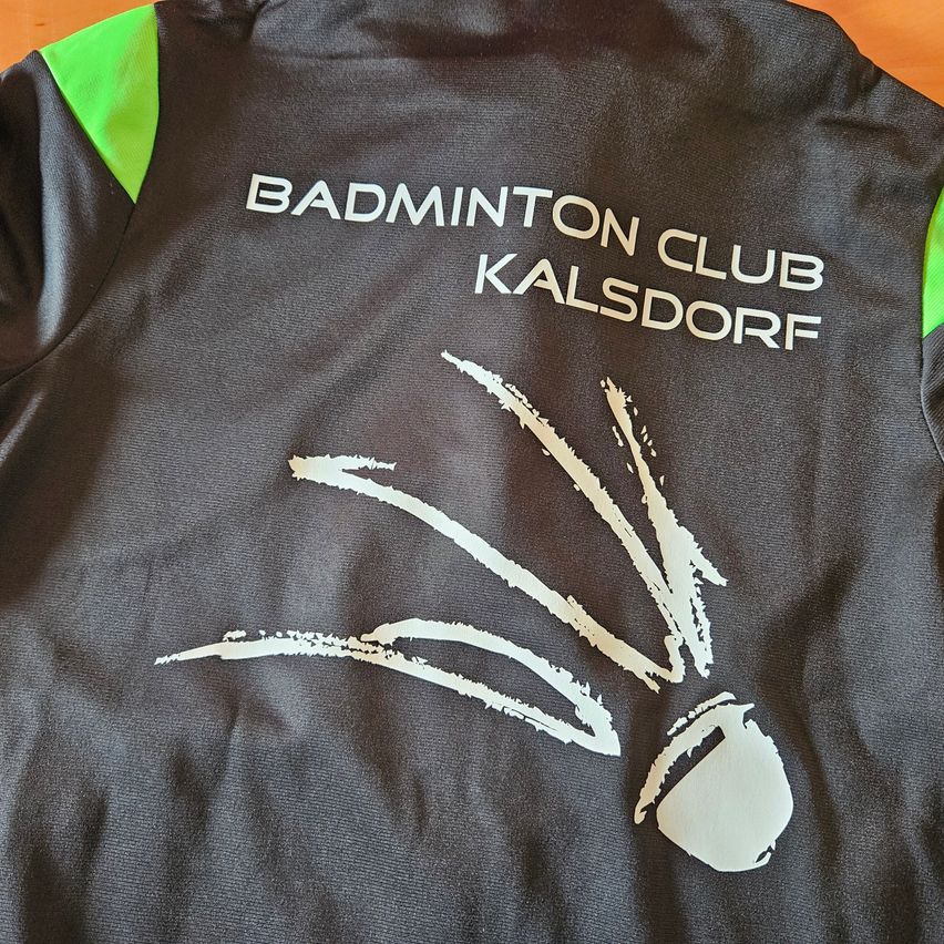 Ein schwarzes Shirt mit weissem Text, der 'BADMINTON CLUB KALSDORF' lautet. Unter dem Text befindet sich eine weisse Illustration eines Badminton-Federballs. Das Shirt hat grüne Akzente an den Ärmeln.
