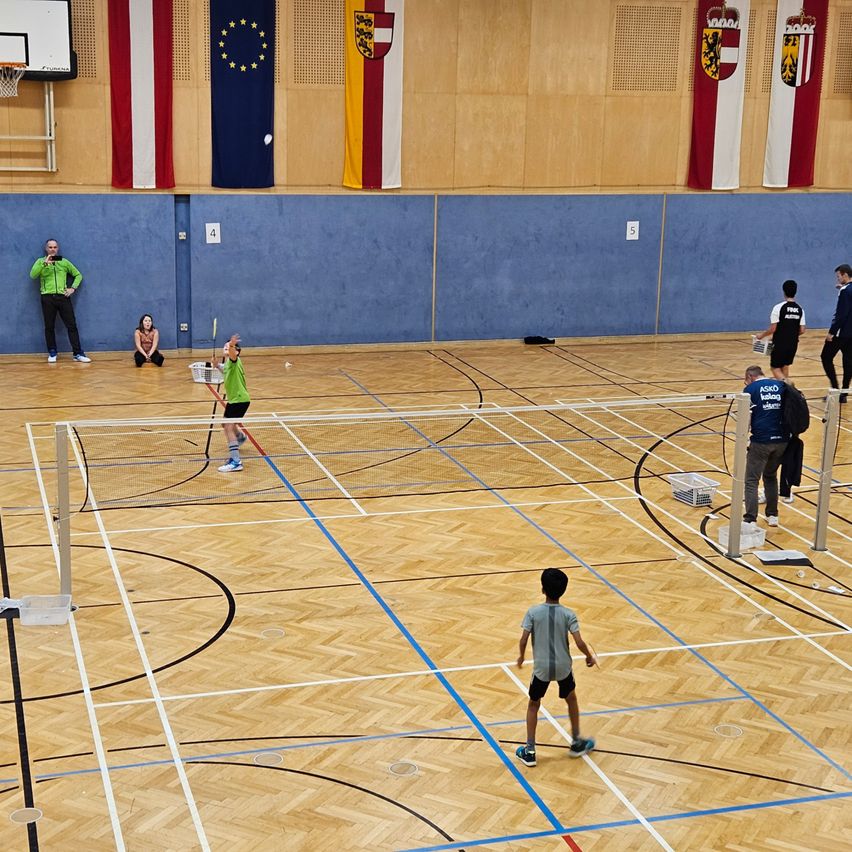 Hallensportplatz mit Holzböden, einige Spieler spielen Badminton, eine Frau sitzt an der Seite, ein Mann steht in der Nähe der Wand, Flaggen hängen an der Wand.