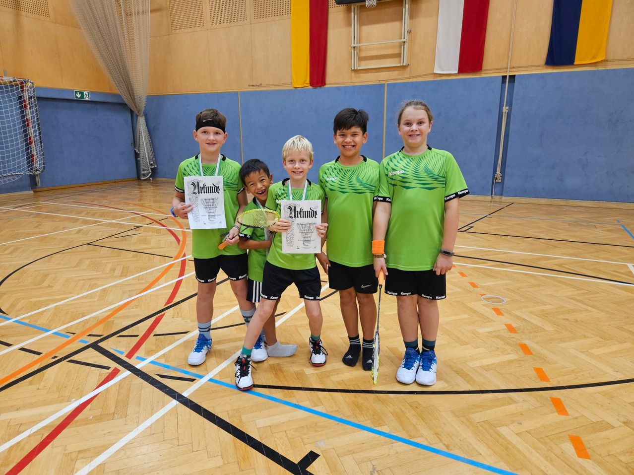 Fünf Kinder in grünen Uniformen und mit Badmintonschlägern stehen in einer Turnhalle und lächeln für ein Foto. Der Hintergrund zeigt eine blaue Wand mit Flaggen und einem Basketballkorb.