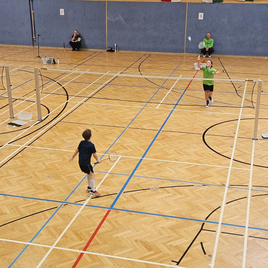 Zwei Personen spielen Badminton in einer Halle, eine springt hoch, um den Federball zu schlagen. Ein Netz trennt sie, und Zuschauer sitzen an der Seite.
