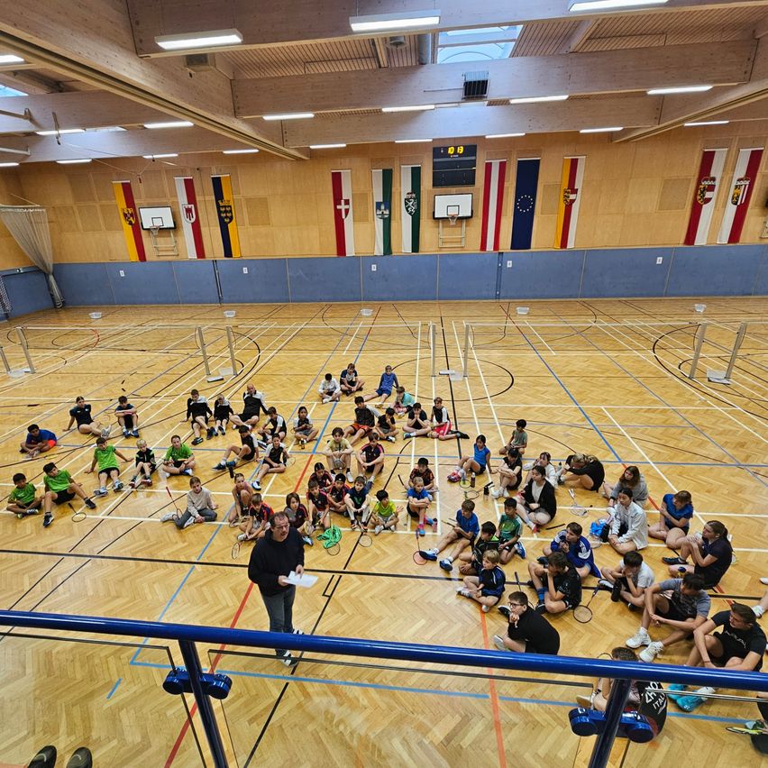 Eine Indoor-Sportanlage mit Holzboden und Fahnen an den Wänden. Eine Gruppe von Menschen sitzt auf dem Boden, möglicherweise eine Badminton-Übung. Ein Mann steht vor ihnen und hält ein Papier in der Hand.
