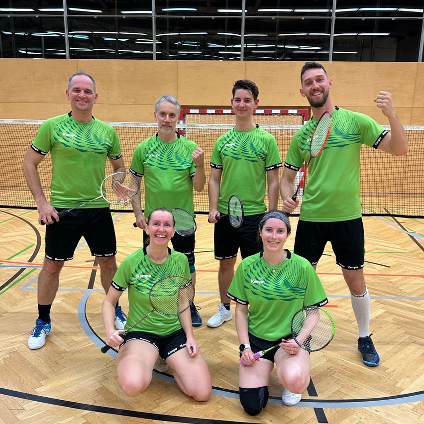 Eine Gruppe von Personen, die grüne Shirts und schwarze Shorts tragen, posiert mit Badmintonschlägern in einer Turnhalle. Zwei Frauen knien, während die anderen stehen. Im Hintergrund befinden sich ein Netz und eine Holzwand.