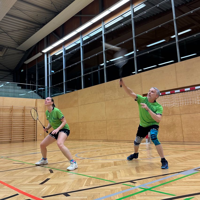 Zwei Personen spielen Badminton in einer Turnhalle mit Holzboden. Einer ist kurz davor, den Shuttlecock zu schlagen, und der andere ist in Bereitschaftsposition.