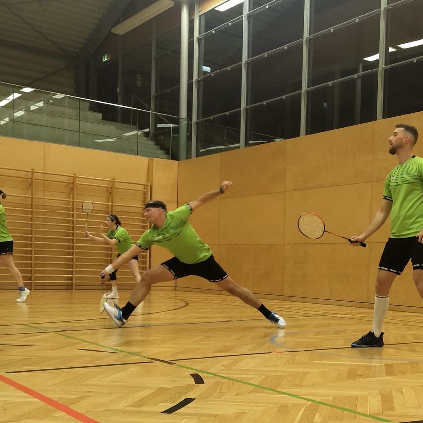 Vier Personen in grünen Shirts und schwarzen Shorts spielen Badminton auf einem Hallenplatz, wobei zwei Spieler in Aktion sind. Der Platz hat Holzboden und braune Wände und Glasgeländer.