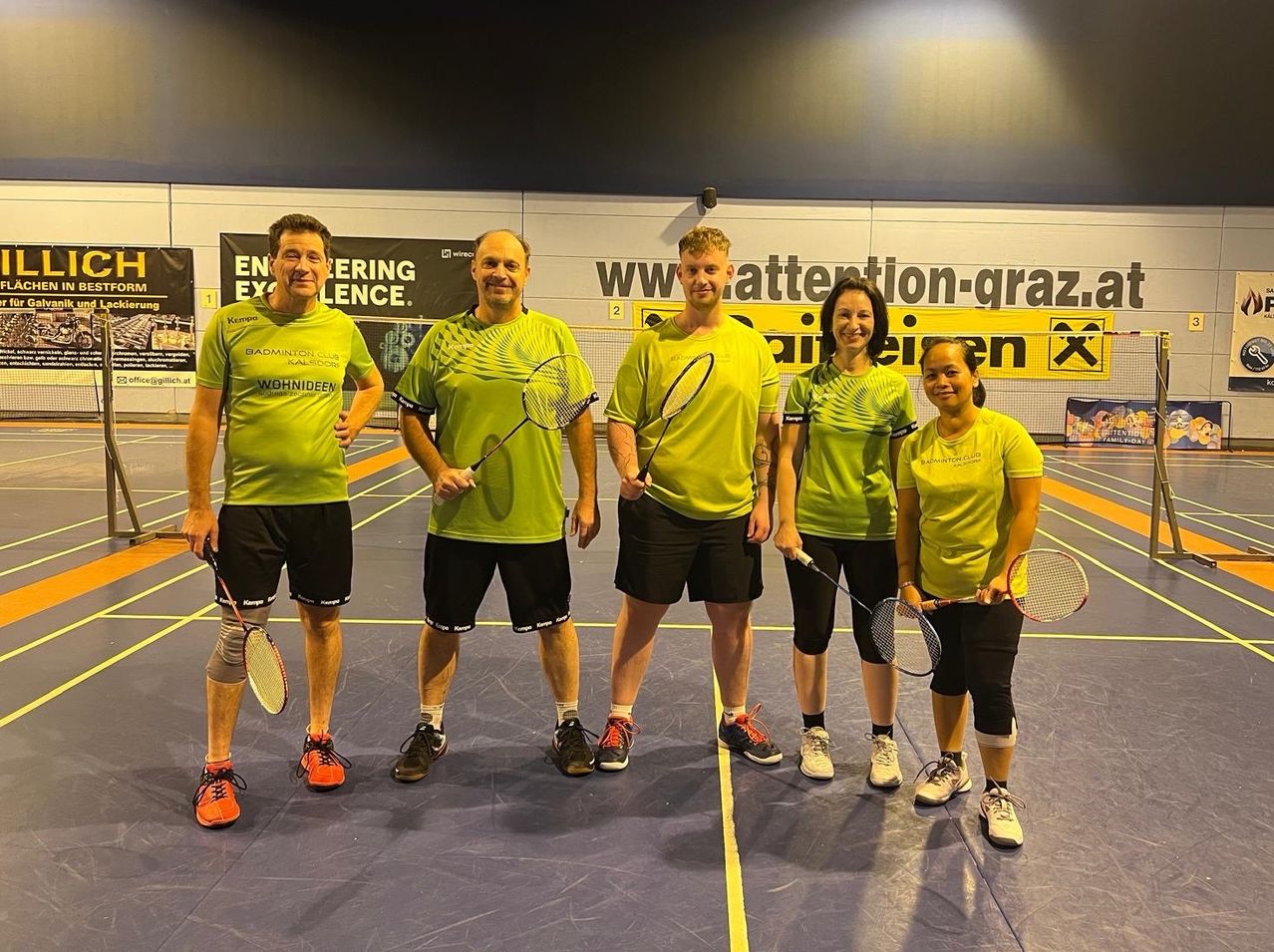Fünf Personen in gelben Trikots posieren für ein Foto auf einem Badminton-Court. Sie halten Schläger und der Court ist von Wänden mit Bannern umgeben.