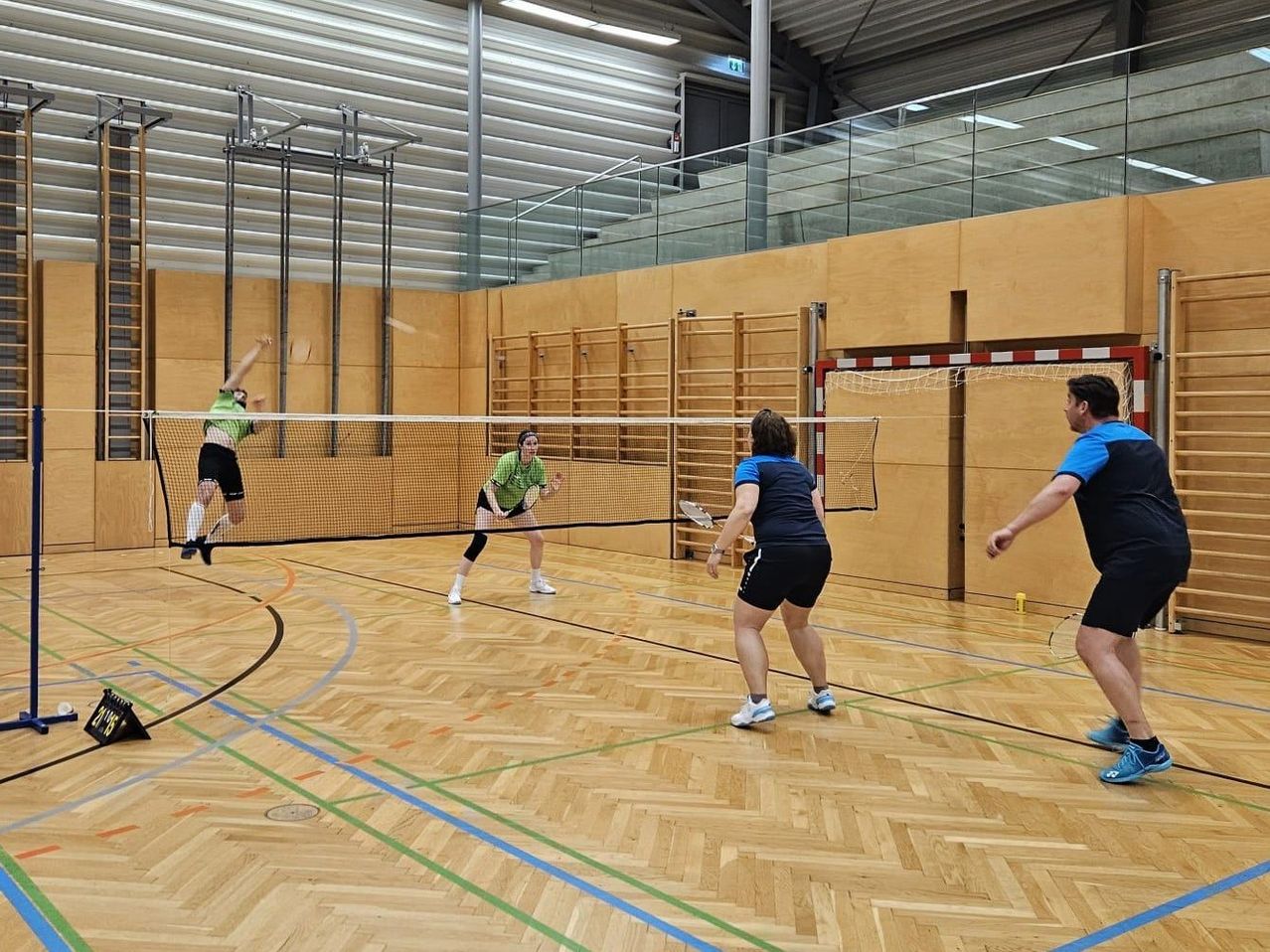 Vier Spieler beteiligen sich an einem Indoor-Badmintonspiel. Zwei Spieler springen für einen Schlag, während zwei andere mit Schlägern stehen, auf einem hölzernen Platz mit einem Netz.
