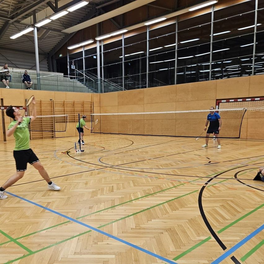 Innen-Badmintonplatz mit aktiven Spielern, Holzböden und hoher Decke mit Beleuchtung. Zuschauer beobachten von den Tribünen.
