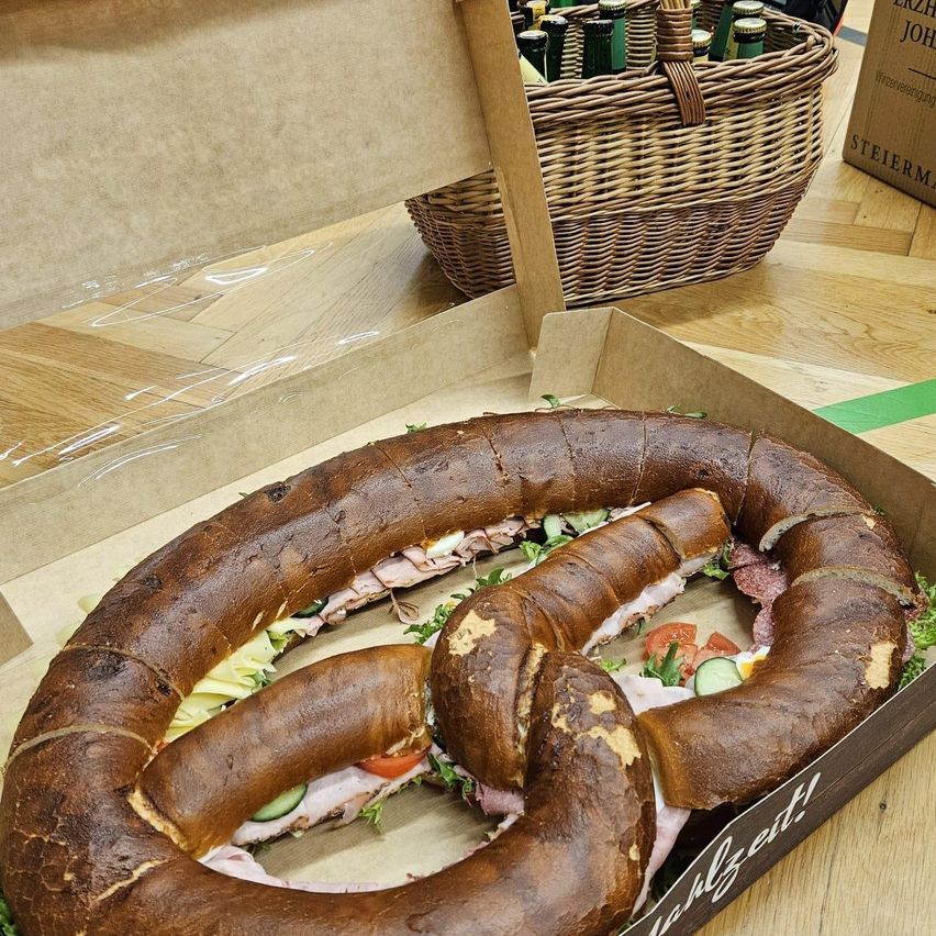 Ein großes Brezelsandwich mit Fleisch, Käse und Gemüse in einer Schachtel, mit einem Korb mit Flaschen dahinter auf einem Holztisch.