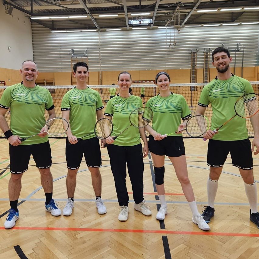 Fünf Personen in grünen Shirts stehen in einer Turnhalle, lächeln und halten Badmintonschläger. Der Boden hat eine hölzerne Oberfläche mit Markierungen. Im Hintergrund ist ein Badmintonnetz zu sehen.