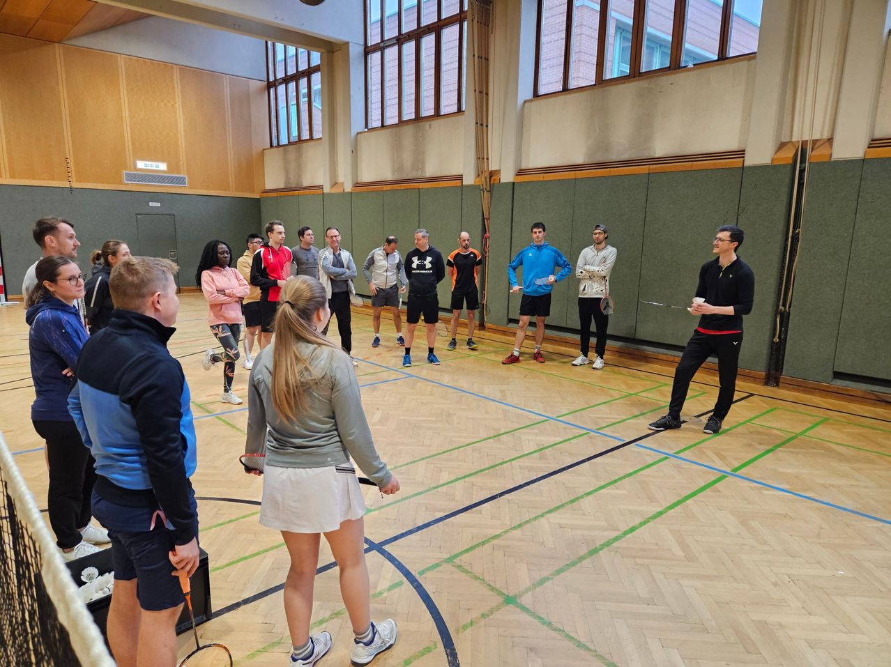 In einer Halle versammeln sich mehrere Menschen in Sportkleidung, einige halten Schläger und andere beobachten. Ein Mann mit Brille spricht die Gruppe an.