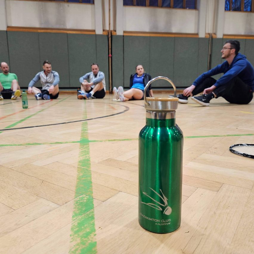 Fünf Personen sitzen auf einem Holzboden, wahrscheinlich in einer Turnhalle. Eine Person trägt eine blaue Jacke, während eine andere eine grüne Flasche hält. Eine grüne Badminton-Flasche steht vor ihnen.