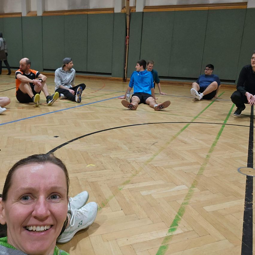 Mehrere Menschen sitzen in einem Kreis auf dem Boden einer Turnhalle und lächeln. Eine Frau vorne macht ein Selfie. Andere tragen Turnschuhe und Freizeitkleidung.