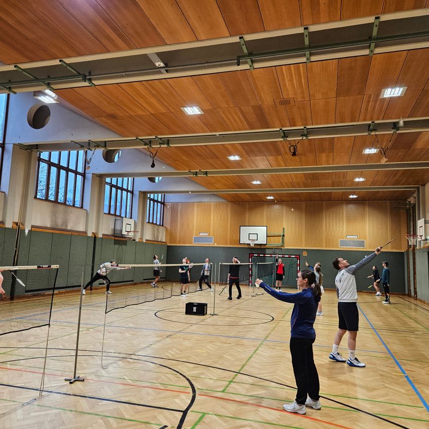 In einer Halle spielen mehrere Personen Badminton mit Schlägern. Sie stehen auf einem Holzboden, der mit verschiedenen Linien und einem Netz markiert ist. Holzbalken und Fenster sind an den Wänden sichtbar.