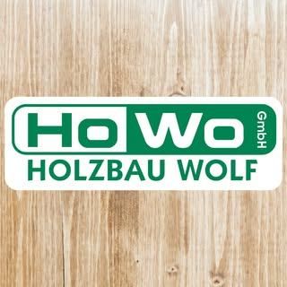 Ein Logo für Holzbau Wolf GmbH auf einer Holzoberfläche. Das Logo zeigt den Firmennamen in grünen Buchstaben auf weißem Hintergrund mit einem grünen Rand. Der Text ist in einer fetten, serifenlosen Schrift geschrieben.