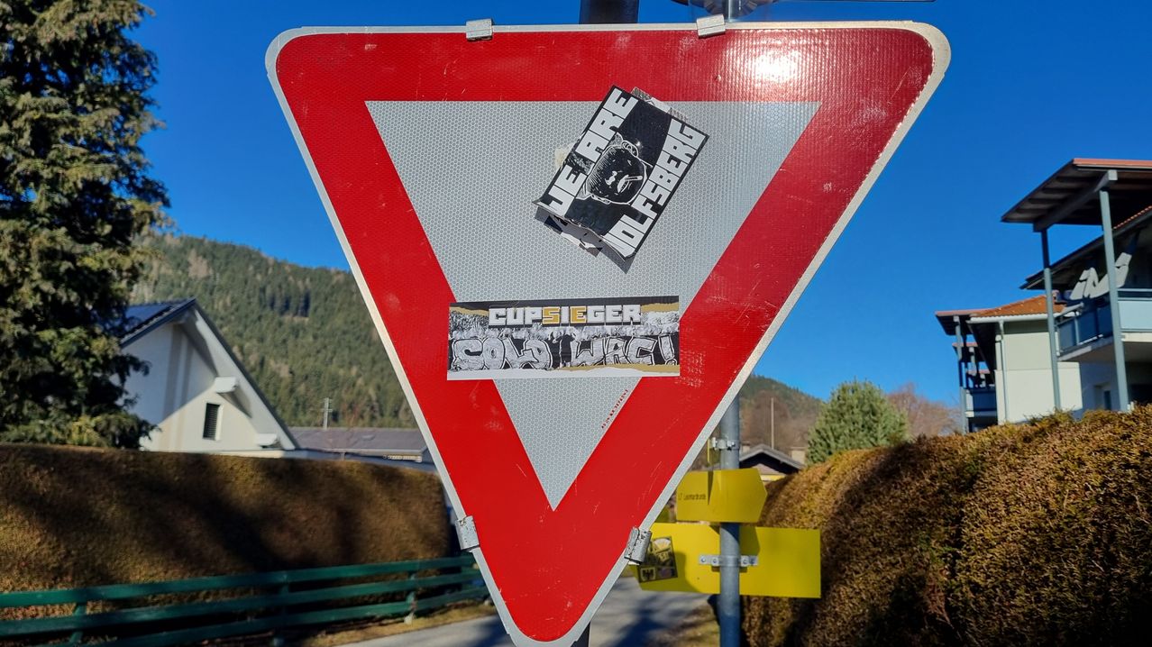 Ein rotes und weißes Vorfahrtsschild mit zwei Aufklebern. Einer lautet 'JE SUIS OLFBERG' und der andere 'CUPS EGER SOLD WACI!'. Dahinter befinden sich Berge und Büsche.