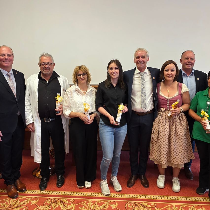 Eine Gruppe von Menschen in formeller und halbformeller Kleidung steht zusammen. Sie halten Auszeichnungen in den Händen und posieren für ein Foto. Der Hintergrund ist schlicht.