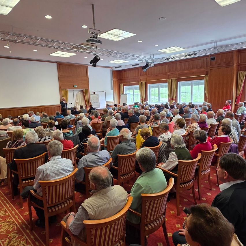 Ein Saal mit Holzwänden und einer weißen Leinwand. Ein Publikum aus überwiegend älteren Menschen sitzt in Stuhlreihen und schaut auf einen Redner auf der Bühne.