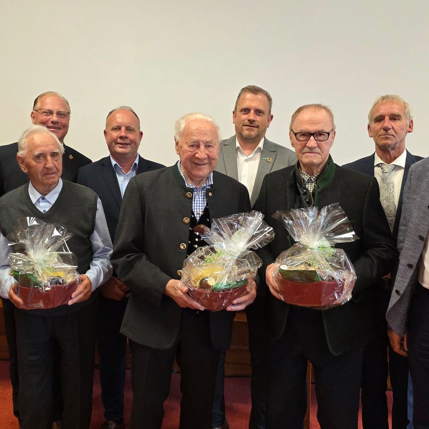 Eine Gruppe älterer Männer steht zusammen und hält Geschenkkörbe. Sie sind formell gekleidet und scheinen einen Anlass zu feiern.