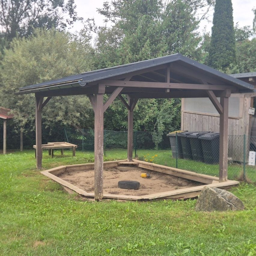 Ein hölzernes Pavillon mit Sandkasten und einem Reifen in einem Grasgarten mit Bäumen im Hintergrund.