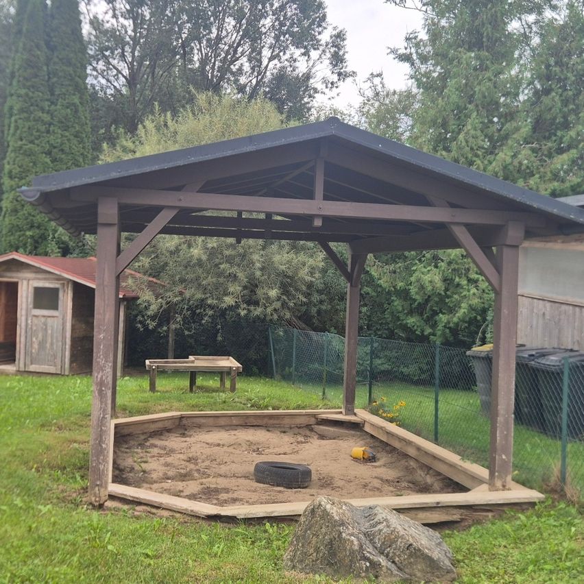 Ein hölzernes Pavillon mit schwarzem Dach steht in einem Garten, umgeben von einem Sandkasten und einem kleinen Holzhaus. Bäume und ein Zaun sind im Hintergrund.