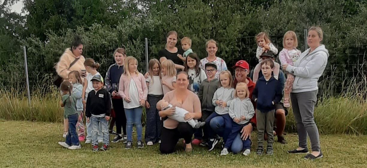 Eine große Familie posiert für ein Gruppenfoto auf einer Wiese, mit Bäumen im Hintergrund. Mehrere Erwachsene halten Kinder, während andere in der Nähe stehen oder hocken.