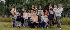 Eine große Familie posiert für ein Gruppenfoto auf einer Wiese, mit Bäumen im Hintergrund. Mehrere Erwachsene halten Kinder, während andere in der Nähe stehen oder hocken.