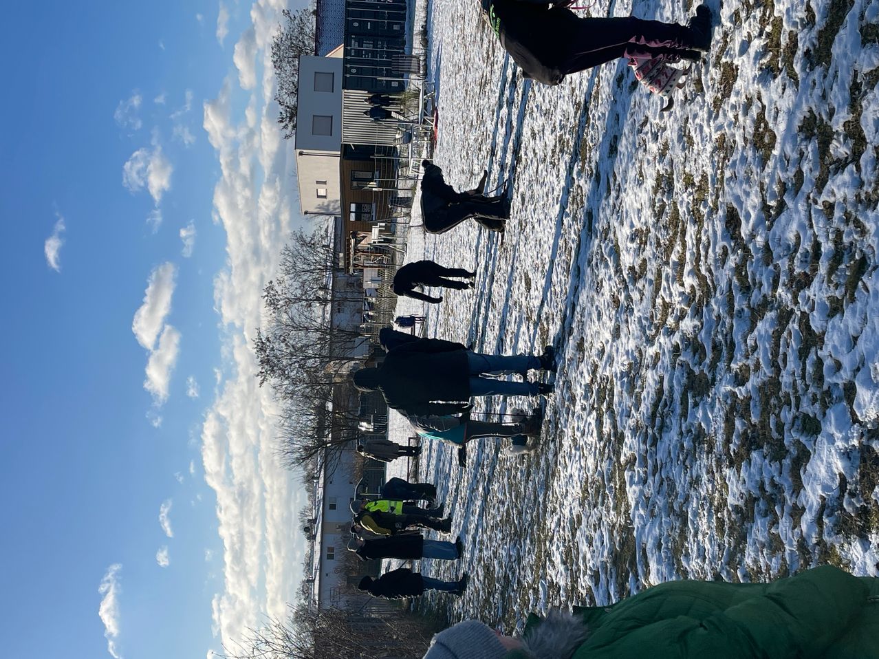 Eine Gruppe von Menschen geht in einem verschneiten Gebiet. Sie tragen Winterkleidung. Einige von ihnen tragen Schuhe, während andere Stiefel tragen. In der Ferne sind Bäume und Gebäude zu sehen. Der Himmel ist blau mit weißen Wolken.