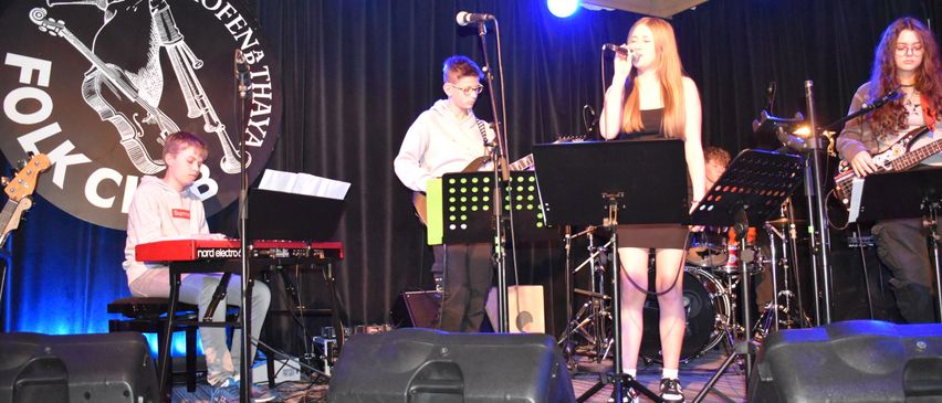 Eine Band tritt auf einer Bühne mit Mikrofon und Musikinstrumenten auf. Ein Mädchen singt und ein Junge spielt Klavier. Ein weiterer Junge spielt Gitarre hinter ihr. Ein Schlagzeug und Lautsprecher stehen auf der Bühne.