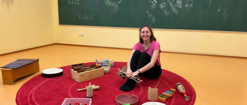 Eine Frau in einem rosa T-Shirt sitzt mit überkreuzten Beinen auf einem roten Teppich, umgeben von verschiedenen Musikinstrumenten. Ein Tafel hinter ihr hat Musiknoten und Instrumentenzeichnungen.