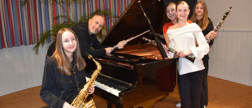 Eine Gruppe junger Musiker posiert für ein Foto mit einem Flügel. Ein Mann hält eine Flöte, eine Frau ein Saxophon und drei Mädchen halten Klarinetten.