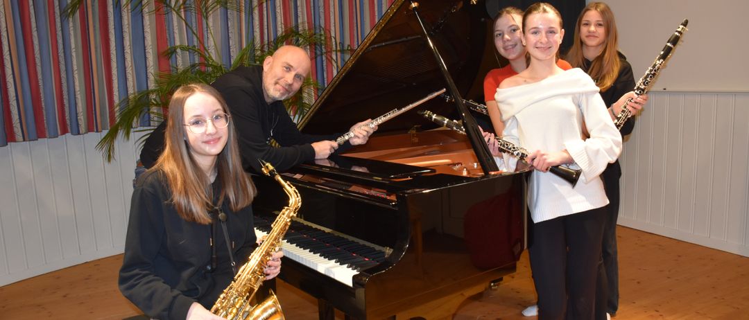 Eine Gruppe junger Musiker posiert für ein Foto mit einem Flügel. Ein Mann hält eine Flöte, eine Frau ein Saxophon und drei Mädchen halten Klarinetten.